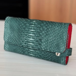 Könnte beinhalten: Eine teal-grüne Geldbörse mit Schlangenmuster. Die Innenseite ist rot und die Geldbörse hat einen silbernen Druckknopfverschluss. Die Geldbörse ist rechteckig und scheint aus Leder zu sein.