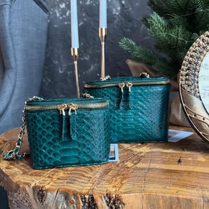 Op de afbeelding: Twee smaragdgroene make-uptassen met gouden ritsen en hardware staan op een houten tafel. De tassen hebben een textuur, slangenleerachtig patroon. Een gouden kettingriem is bevestigd aan de kleinere tas.