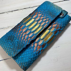 May include: A blue, gold, and copper-coloured wallet with a scale-like pattern. The wallet has a snap closure and is made of a textured material. The design is reminiscent of a peacock's feathers.