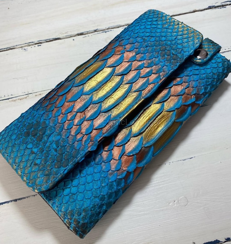 K&ouml;nnte beinhalten: Eine blaue, gold- und kupferfarbene Geldb&ouml;rse mit schuppenartigem Muster. Die Geldb&ouml;rse hat einen Druckknopfverschluss und ist aus einem strukturierten Material gefertigt. Das Design erinnert an Pfauenfedern.