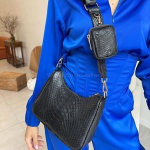 May include: Black leather shoulder bag with a small matching pouch attached to the strap. The bag has a textured surface and a rectangular shape with rounded corners. The strap is adjustable and features gold-coloured hardware.