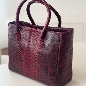 Op de afbeelding: Een bordeauxrode tote bag met een slangenpatroon. De tas heeft twee afgeronde handvatten en een rechthoekige vorm. Het slangenpatroon is een mix van bordeauxrood en zwart, waardoor de tas een textuur krijgt. De tas staat op een wit oppervlak.