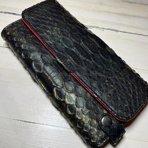 May include: Black leather wallet with a reptile scale pattern and gold detailing. The wallet features a red trim and a snap closure. The wallet is on a white wooden surface.