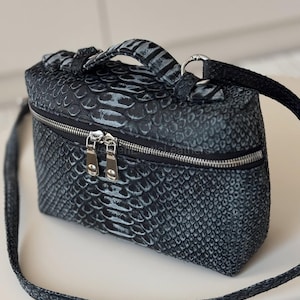 Op de afbeelding: Een kleine, rechthoekige zwarte en grijze cosmetische tas met een textuur, slangenleerpatroon. De tas heeft een zilveren ritssluiting, een handvat aan de bovenkant en een afneembare schouderriem. Ontworpen voor make-up en andere schoonheidsproducten.