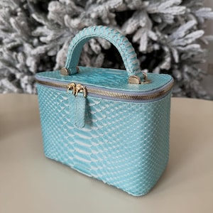 Op de afbeelding: Een kleine, rechthoekige, turquoise handtas met een handvat aan de bovenkant en een gouden rits. De tas heeft een textuur met schubbenpatroon en gouden hardware. De handgreep is ook turquoise en heeft een gevlochten ontwerp.