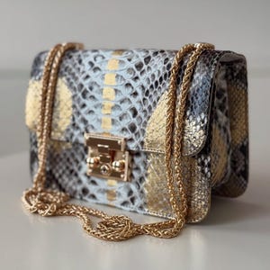 Op de afbeelding: Een kleine, rechthoekige handtas met een slangenprint in blauw, grijs en goud. De tas heeft een gouden kettingriem en een gouden sluiting. Een stijlvol accessoire.