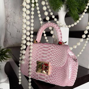 May include: A pink handbag with a snakeskin texture. The bag features a gold clasp and handle. White pearl beads and green pine branches are in the background.