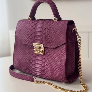 Może przedstawiać: Śliwkowa torebka z teksturowanym wzorem skóry węża. Torebka posiada górny uchwyt, klapę z pozłacanym zapięciem i złoty łańcuszek. Torebka jest w stylu strukturalnej aktówki.