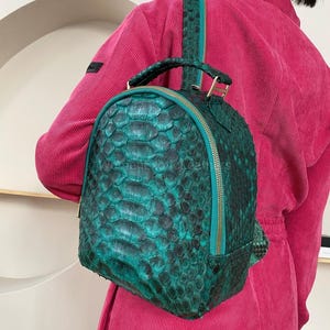 Puede incluir: Una mochila pequeña con un patrón texturizado similar a escamas en tonos verde esmeralda intenso y negro. La bolsa tiene una parte superior redondeada, un asa superior y una cremallera color verde azulado que recorre el perímetro.