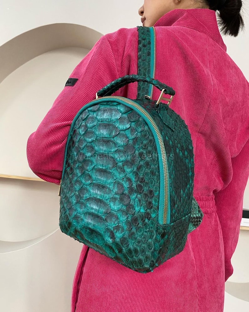 Op de afbeelding: Een kleine rugzak met een gestructureerd, schubachtig patroon in tinten diep smaragdgroen en zwart. De tas heeft een afgeronde bovenkant, een handvat aan de bovenzijde en een blauwgroene ritssluiting die langs de omtrek loopt.