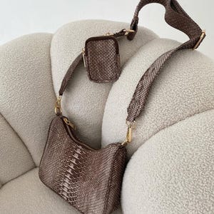 May include: A brown and beige snakeskin-effect shoulder bag with a matching small pouch. The bag features a gold zip and hardware, and a wide, adjustable strap. The bag is on a white, textured surface.