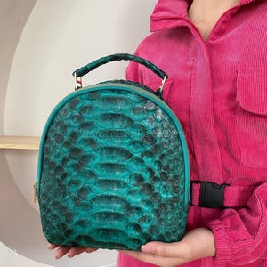 Może przedstawiać: Turkusowy plecak z teksturowanym wzorem przypominającym łuski. Torba ma zaokrąglony kształt, górny uchwyt i złoty zamek błyskawiczny. Słowo "HONEY" jest wytłoczone z przodu. Torba jest trzymana na różowym tle.