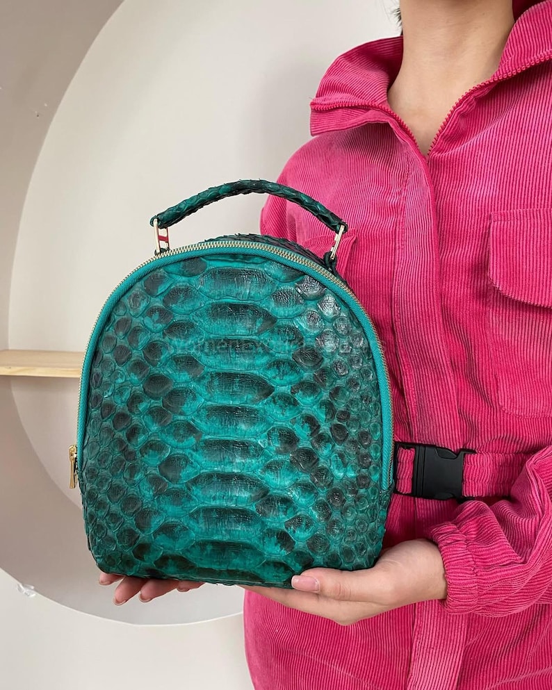 Op de afbeelding: Een afgeronde rugzak met een gestructureerd slangenleerpatroon in groenblauw en zwart. De tas heeft een handvat aan de bovenkant met een gevlochten ontwerp, een goudkleurige ritssluiting en een bijpassende groenblauwe rand langs de randen.