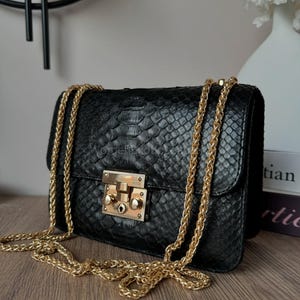 Op de afbeelding: Zwarte leren handtas met een slangenleertextuur, met een goudkleurige kettingriem en sluiting. De tas is rechthoekig met een klepsluiting. De ketting is over de tas gedrapeerd. De tas staat op een houten oppervlak.