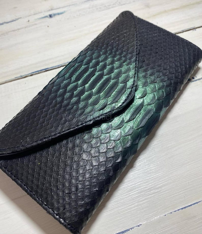Puede incluir: Una cartera de cuero negro con textura de piel de serpiente y cierre de solapa. La cartera tiene un degradado de tonos verdes y negros. La cartera es rectangular y parece ser de cuero genuino.