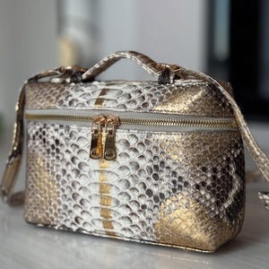 May include: A rectangular, gold, white, and brown snakeskin-patterned bag with a top handle and a shoulder strap. The bag features a silver zipper with gold-coloured double pulls. The bag is approximately 20 cm long, 15 cm wide, and 13 cm tall.