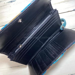 K&ouml;nnte beinhalten: Ge&ouml;ffnete schwarze Ledergeldb&ouml;rse mit mehreren Kartenf&auml;chern und einem Rei&szlig;verschlussfach. Die Au&szlig;enseite weist ein farbenfrohes Muster mit Blau-, Gelb- und Oranget&ouml;nen auf. Die Geldb&ouml;rse ist ge&ouml;ffnet und zeigt die Innenf&auml;cher.