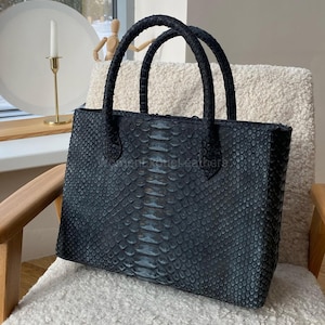 Könnte beinhalten: Eine schwarze Leder-Tragetasche mit Schlangenmuster. Die Tasche hat zwei abgerundete Griffe und eine rechteckige Form. Die Tasche steht auf einem weißen, flauschigen Stuhl. Der Hintergrund umfasst ein Fenster, einen Kerzenhalter und eine Holzfigur.