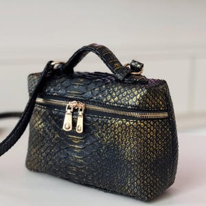 May include: A small, black and gold faux snakeskin handbag with a top handle and a detachable shoulder strap. The rectangular bag has a double zip closure and gold-toned hardware. The bag's textured surface has a scale-like pattern.
