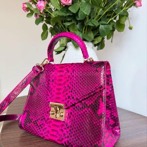 May include: A bright pink handbag with a snakeskin design. The bag has a top handle, a detachable shoulder strap, and a gold-tone clasp. The background features pink roses in a white vase, creating a striking contrast.