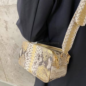 May include: A rectangular crossbody bag with a snakeskin pattern in shades of gold, beige, and grey. The bag features a gold zip closure and a matching adjustable strap. The text "ExoticLeathers" is visible on the bag.