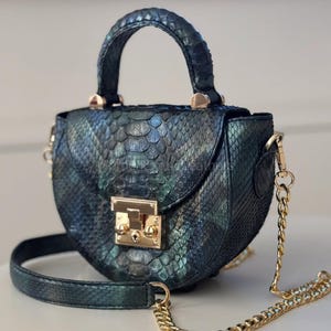 Op de afbeelding: Een kleine, ronde handtas met een getextureerde, iriserende afwerking in tinten van teal en zwart. De tas heeft een handvat aan de bovenkant en een gouden kettingriem. Een gouden sluiting sluit de voorklep af. Afmetingen: ongeveer 18 cm breed en 15 cm hoog.