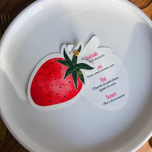 Peut inclure: Une carte de menu en forme de fraise avec une illustration de fruit rouge et des feuilles vertes, fixée par une attache dorée sur une carte blanche contenant du texte français dont Cocktails, Entrée, Plat et Dessert, posée sur une assiette.