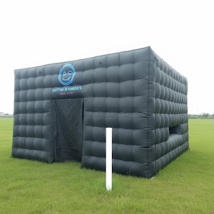 Pode incluir: Uma grande estrutura insuflável preta com um design quadrado e acolchoado. A estrutura tem uma entrada com uma cortina preta e uma janela. O texto "CHITTRA & VANDA'S RAVE CAVE" é exibido acima da entrada. A estrutura está num campo relvado.