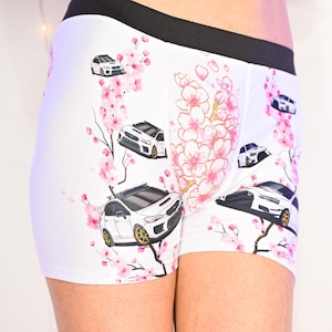 May include: White boxer briefs featuring a repeating pattern of white sports cars with gold wheels and pink cherry blossom branches. The garment has a wide black elastic waistband and a fitted silhouette with a graphic print design.