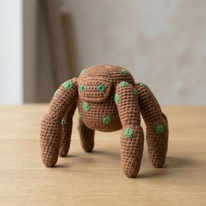 May include: A handmade brown crochet spider toy with six legs and a rounded body. The spider has green polka dots on its body and legs. The toy is sitting on a wooden surface.