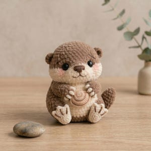 May include: A crocheted otter figurine in shades of brown and cream, holding a small, tan crocheted shell. The otter has rosy cheeks, black eyes, and a small tail. The figurine is sitting on a wooden surface.
