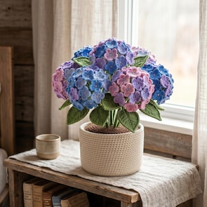 May include: A crocheted hydrangea plant with blue, purple, and pink flowers in a beige pot. The handmade flowers have green leaves and are displayed on a wooden table with a small ceramic cup.