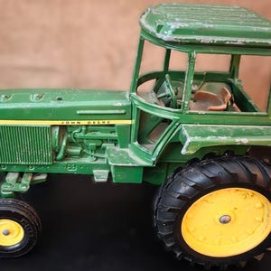 May include: Green John Deere toy tractor with yellow accents on the wheels and a yellow stripe. The tractor has a cab with a steering wheel and a seat. The words "JOHN DEERE" are printed on the side.
