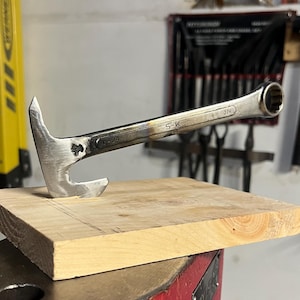 May include: A custom-made metal tool fashioned from a silver-toned S-K brand wrench, featuring a pointed claw-like head and a box-end handle. The tool is embedded into a square piece of light-colored wood, which rests atop a red metal anvil base.