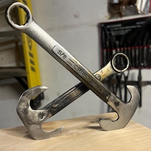 May include: A metal anchor sculpture crafted from two crossed wrenches. The longer wrench is marked with the text 5/8 CRAFTSMAN, while the shorter wrench features the number 12. The silver-toned metal has a weathered, industrial finish.