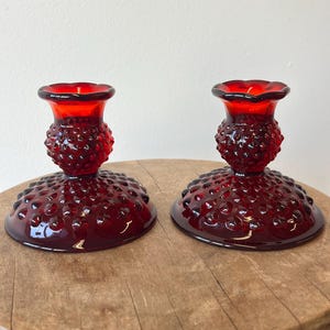 Puede incluir: Dos candelabros de vidrio rojo rubí. Cada uno tiene una base redonda cubierta de pequeñas protuberancias, un tallo texturizado y una parte superior acampanada. Los candelabros se exhiben sobre una superficie de madera sobre un fondo blanco. El vidrio tiene un acabado brillante.