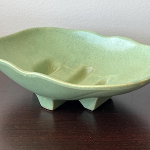 Puede incluir: Un jabonera de cerámica verde claro con una textura moteada. El plato tiene una forma única, festoneada y descansa sobre tres pequeños pies triangulares. El interior presenta crestas elevadas para el drenaje del jabón. Un accesorio de baño vintage.