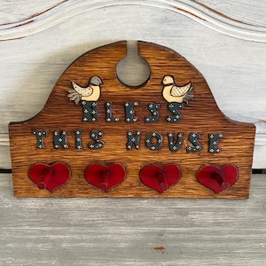 Op de afbeelding: Houten wandophanging met de woorden "BLESS THIS HOUSE" en twee vogel figuren. De bruine plaat heeft vier rode hartvormige haken. De vogels zijn beige en grijs.