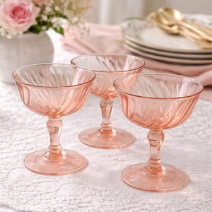 May include: Three pink-tinted coupe glasses with a spiral design. The glasses are on a white lace tablecloth, with a vase of pink roses in the background. The glasses have a classic, elegant design.
