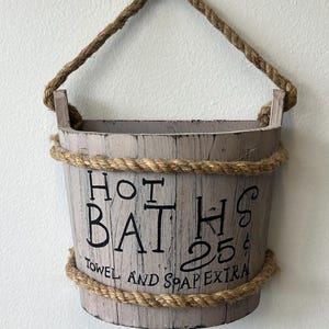May include: A rustic, wooden bucket with a rope handle and trim. The bucket is painted in a distressed, light gray color and features the words "HOT BATHS" in black script, along with "TOWEL AND SOAP EXTRA".