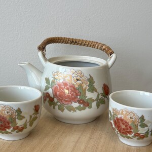May include: A cream ceramic tea set comprising a teapot with a woven handle and two cups. The set is adorned with a floral pattern in red, orange, and green, offering a vintage appeal. The teapot and cups are ideal for serving tea.