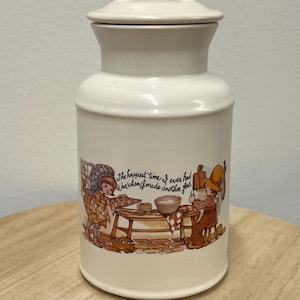 Pode incluir: Pote de cerâmica creme com tampa e alça. O pote apresenta uma ilustração colorida de crianças a cozinhar, com o texto "The happiest time I ever had was when I made another glad."