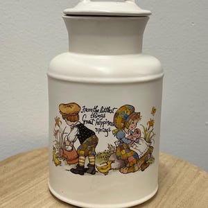 Pode incluir: Uma caneca de cerâmica branca com uma alça preta e o texto "But First, Coffee" em negrito preto. A caneca está sobre um fundo desfocado, sugerindo um ambiente acolhedor.