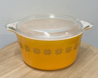 May include: A vintage, round, yellow Pyrex bowl with a clear glass lid. The bowl features a repeating starburst pattern in a darker yellow color. The bowl is sitting on a light brown wooden surface.