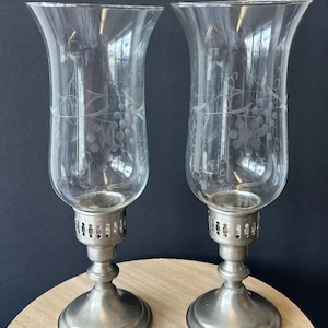 May include: Two clear glass candle holders with etched floral designs. The holders have a flared top and sit on a silver-colored metal base. The bases are round with a decorative design. The candle holders are displayed on a wooden surface.