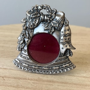 May include: A silver-coloured bell-shaped picture frame with a red velvet backing. The frame is decorated with a bow, holly, and berries. The frame is approximately 7.6 cm tall and 6.3 cm wide.