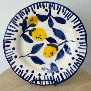 May include: White ceramic plate with a blue rim and a hand-painted lemon design. The plate features yellow lemons and blue leaves on a white background. The rim has a blue dotted and striped pattern.