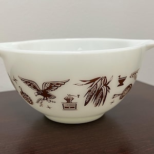 Puede incluir: Cuenco mezclador Pyrex blanquecino con ilustraciones decorativas marrones. El cuenco presenta imágenes de un águila, maíz y un molinillo de café. Tiene dos asas y es un artículo de cocina clásico.