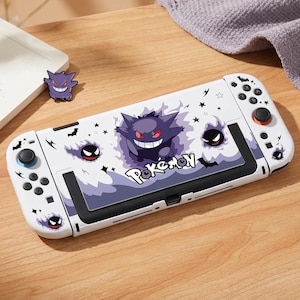 Può includere: Custodia bianca per console Nintendo Switch con un design Gengar viola e la scritta "Pokemon". La custodia ha pulsanti neri e uno schermo nero. È presente anche una spilla Gengar viola.