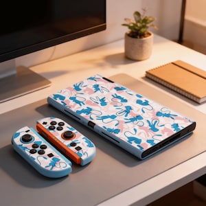 Peut inclure: Une console Nintendo Switch bleu clair et des manettes avec un motif répétitif de personnages de dessins animés bleus et roses. Les manettes ont des boutons noirs et des accents orange. La console et les manettes sont sur une surface grise.
